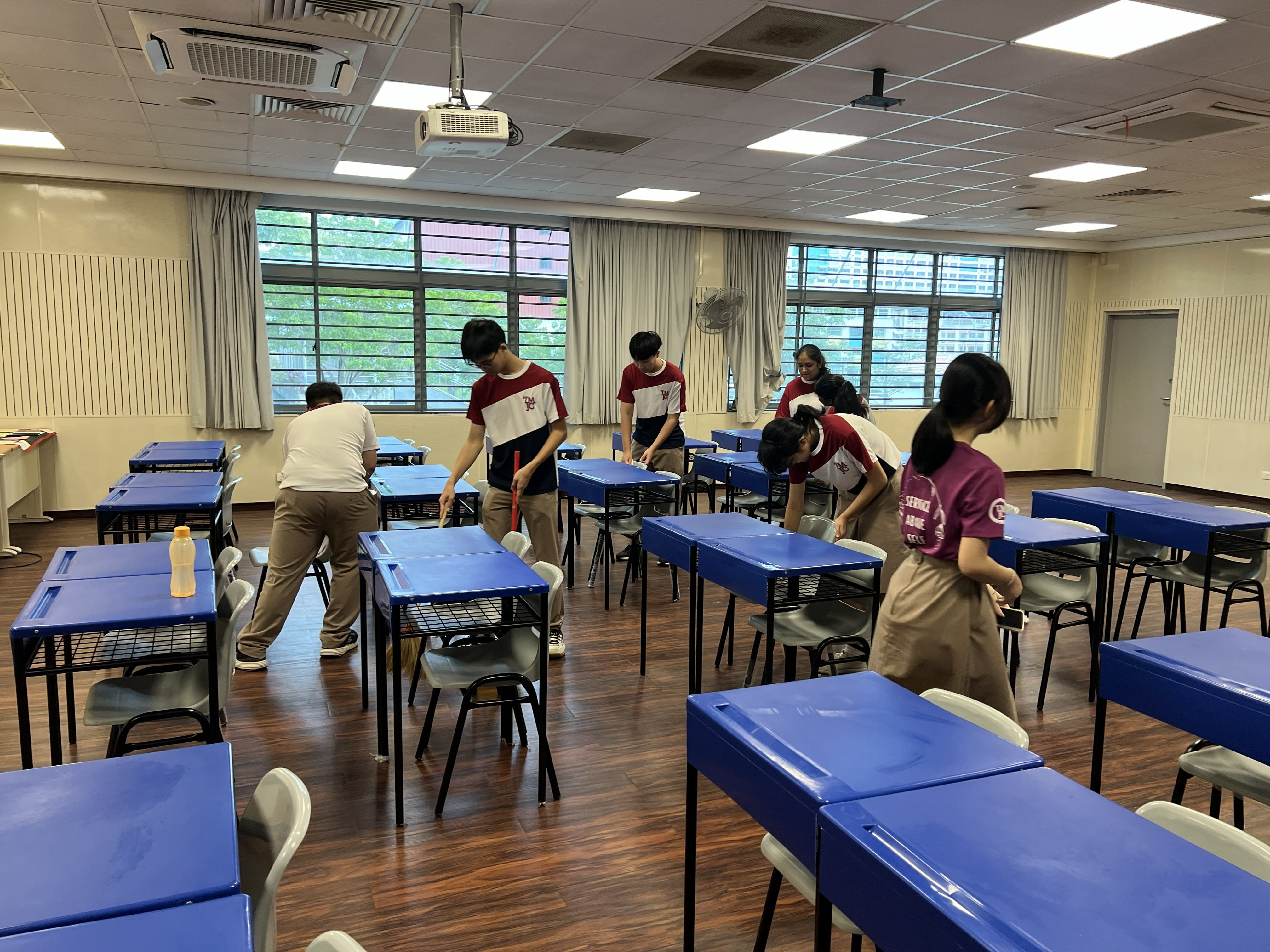 A group of students in TMJC uniforms clean a classroom together ahead of Chinese New Year, sweeping floors and wiping down blue desks. One student's shirt reads "Service Above Self."